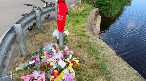 A picture of a pole next to the canal that is surrounded by flowers on the floor. Attached to the pole is a red signed football shirt.