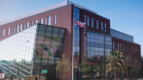 The Castle House building is a modern structure, with big windows and trees around it. There is also a union flag flying outside. 