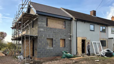 Luke Deal/BBC A photo taken outside where a house renovation is taking place. A new house build can be seen attached to already constructed homes. It is made of large grey bricks and has scaffolding on one side of it. The windows are covered in wooden planks. A man stands in the doorframe of part of the house. 