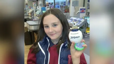 Joshua McParland Joshua as a young boy. He wears his hair long and is smiling at the camera. He is wearing a red sweater and a blue body warmer.  He is holding a small white ball which has his name written on it. The photo is taken beside the checkout in a small shop. 