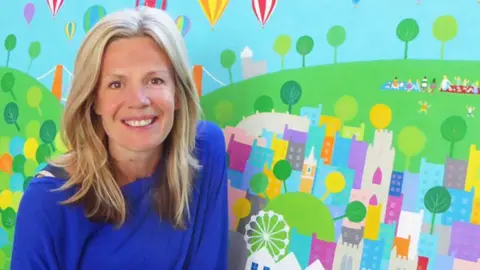 Wearing a blue top, Jenny smiles at the camera whilst posing in front of one of her artworks. She has blonde hair hanging loose below her shoulders. The art depicts hot air balloons flying over Bristol. The Clifton Suspension Bridge can be seen in the work behind her, as can children playing in a park. 