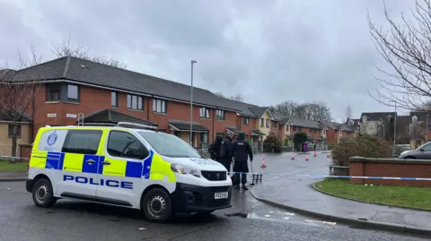 Police officers and a police van at the entrance to a cul de sac of several redbrick homes, with gardens and cars parked in front of the homes.