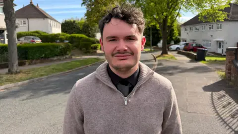 A man with curly hair and a grey fleece top is facing the camera and standing in a tree lined street