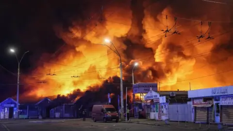 Large fire engulfs a market area at night, sending towering flames and thick smoke into the sky above nearby buildings, streets, and overhead power lines.