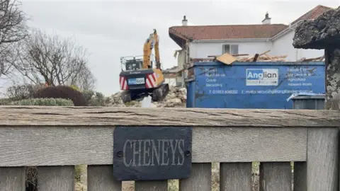 John Fairhall/BBC A digger in the distance on top of a mound as it demolishes a house. The house is white with a red roof. The entrance gate into the house is at the bottom of the picture with the name "Cheneys" on a black slate on the fence.