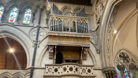 The interior of a large historic church with an ornate pipe organ mounted high on the wall. The casing is decorated with Gothic-style tracery, gold accents, and painted motifs.