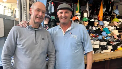 Michael on the left has a grey long sleeved top on and is bald. Next to him is Gil who has a flat cap on and a light blue t-shirt. He has his arm around Michael and in the background is a market stall full of hats on mannequin heads and scarves hanging up