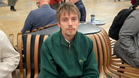 Josh Wooton sitting a wooden slat bench in a train station. He is wearing a green jacket and has short blonde hair and wispy beard. 