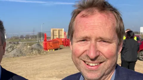 Luke Deal/BBC Richard Winch on the site where the new innovation centre will be building. He wears a navy suit jacket with a blue shirt underneath. 
