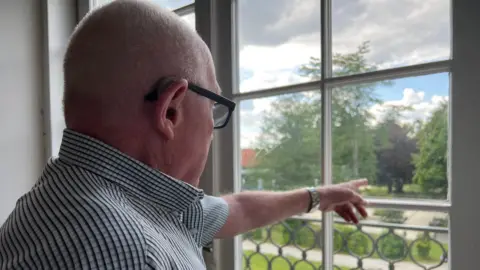 Roy has short hair and glasses, and is wearing a striped shirt, stands indoors and points outside while looking through a window. Outside the window, there is a green lawn, trees, and a partly cloudy sky."
