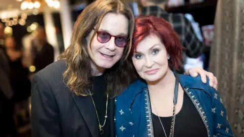 Ozzy Osbourne and Sharon Osbourne. He has long brown hair and is wearing a black jacket and glasses and Sharon is wearing a blue jacket and has redish brown hair. 