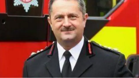 NIFRS Gary Thompson pictured in NIFRS uniform, black suit with red lapels, white shirt and black tie. Thompson has short grey hair and a moustache. He is standing in front of a fire engine, which is red and yellow. 