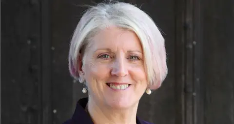 Dr Lynne Sedgmore Dr Lynne Sedgmore CBE smiling at the camera wearing pearl earrings and white hair with a pink of pink die