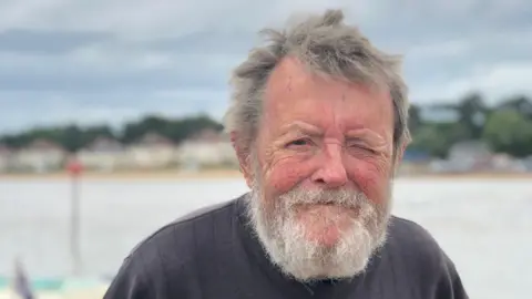 Holly Brookes A man with grey hair and a grey beard smiles at the camera. He wears a grey jumper and he is standing in front of a river. 