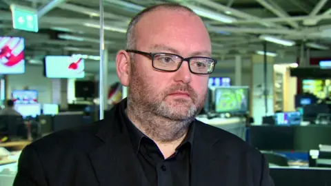 A man is standing in an office. He's wearing glasses with a black frame and a black shirt and jacket. There are a lot of TVs in the background.