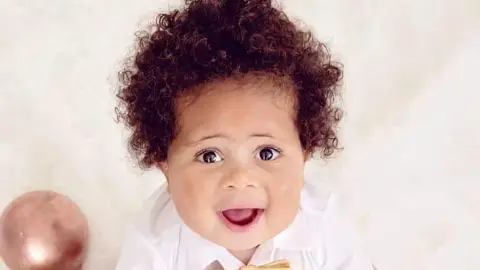 The image shows a young infant sitting upright on a soft, white, fluffy surface that looks like a studio backdrop or blanket. The infant is facing the camera, with the head slightly raised and both hands placed on the surface for support. The infant is wearing a white long-sleeved shirt with a small light-coloured bow tie at the collar.
