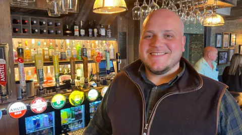 Rob Moore is standing at a bar with all the measures and beer taps in the background. He is wearing a blue and yellow checked shirt with a brown zip-up gilet over the top and smiling at the camera.
