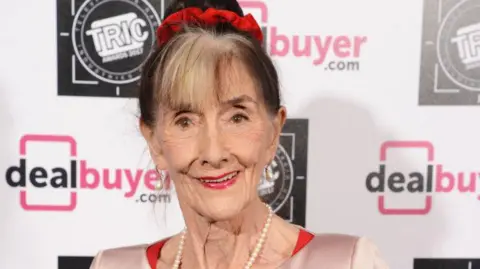 Getty Images A close-up of June Brown as she smiles and looks into the camera. She has her hair tied up in a bun and is wearing pearl drop earrings and a pearl necklace