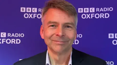 Andrew Gant, who is wearing a collared shirt and jacket, smiles at the camera in front of a blue background bearing the logo of BBC Radio Oxford.