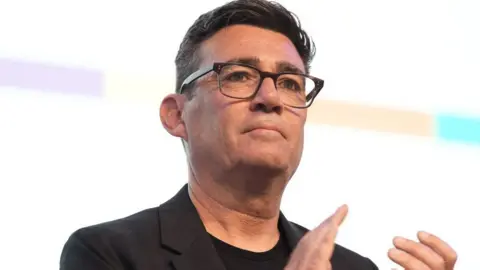 Andy Burnham, who has short black hair and glasses, in a black suit jacket. 