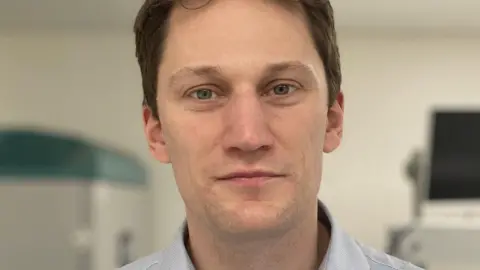 CUH A close-up of Matthew Smith, who has short light brown hair and is wearing a fine stripped blue and white shirt. He is standing in a white room.