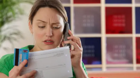 A woman on the phone wearing a green top. She looks upset and is holding a credit card and a paper bill. She is sitting in front of a book shelf and plant.