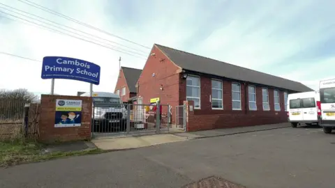 An image of Cambois Primary School with its sign in blue with white letters. The building is brown brick and there is a gate with a mini bus parked behind it. 