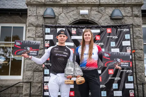 Pete Scullion Two bike riders - one man and one woman - hold up trophies and signs saying Beijing, China. The man has a black baseball cap on back to front with a black top and white bottoms. The woman has a Slovakia top on and black bottoms.