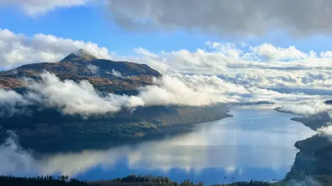 A mountain beside a loch, with low clouds hanging over the water and reflections 