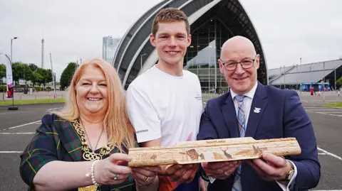 Three people holding a wooden baton in front of the Armadillo venue in Glasgow. From left to right is a woman with long blonde hair, wearing a tartan blazer and an ornate gold chain around her neck. Next to her is a tall young man wearing a white t-shirt. Next to him is a bald man with glasses wearing a blue blazer, purple shirt and blue checked tie.