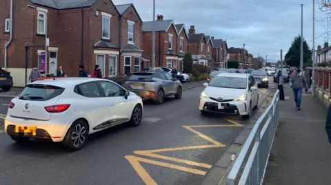 Nine cars on the road outside Netherfield Primary school. A white car is parked on the yellow zigzags which is creating a traffic jam.