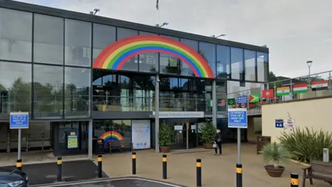 Google A three-floor glass hospital building with a rainbow painted on the outer windows of the top floor. There are flags on the walkway to the right