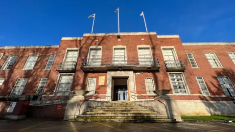 BBC Looking up at Swindon's civic offices under a bright blue sky. Steps up to the front of the red brick building with three flag poles on top.
