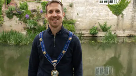 Bradford on Avon Town Council A man in a mayoral chain stands in front of a river and brick building.