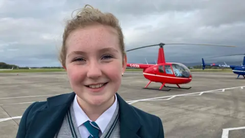 Jessica wears her school uniform -a teal jacket with grey jumper, teal stripe round the collar and teal tie. She has blonde hair tied up