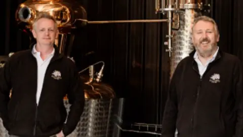 Jim Nash Shows two men in dark zip-up tops and white shirts standing in their distillery