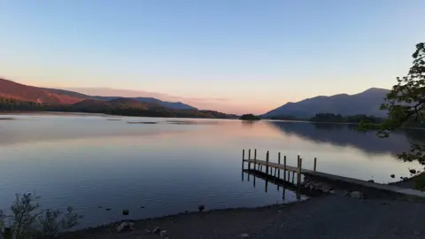 The Field House Fairy A large lake with a wooden jetty in the foreground. The lake is partly in shadow on the right hand side. The sky is light blue with an orangey glow towards the horizon. There are mountains in the background with trees surrounding the lake.