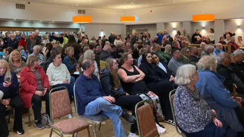 A large gathering of people sitting on chairs in a hall with others standing at the back