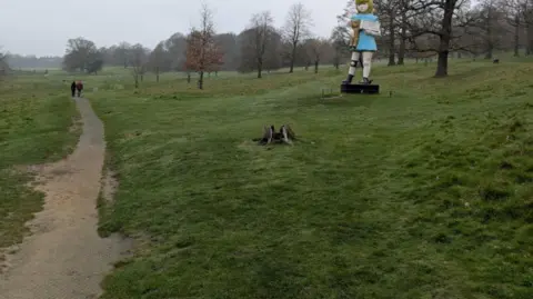 David Spereall/BBC A large sculpture of a girl holding a teddy. It stands in a grassed area with trees dotted around. Two people walk on the footpath ahead.