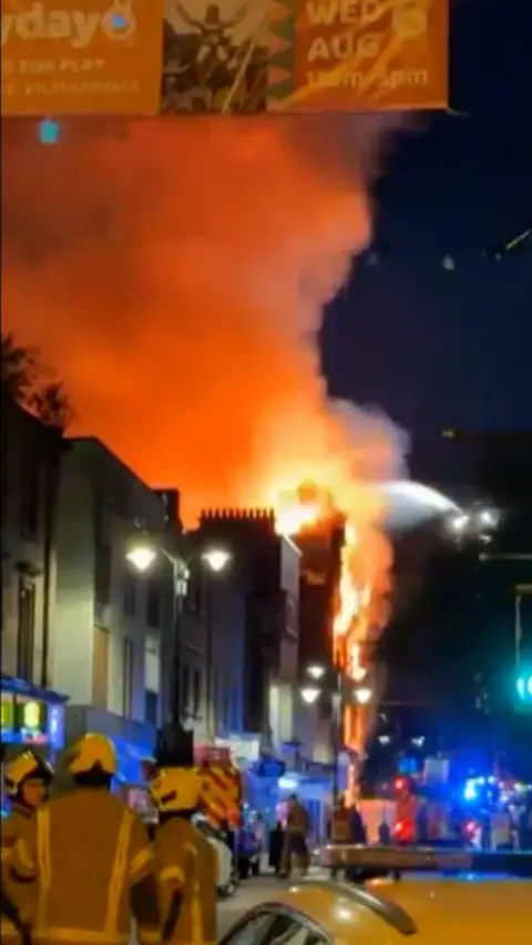 Firefighters look at flames and smoke coming out the top of a building