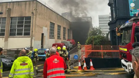 Black smoke billows our of Royal Mail depot in Brighton and firefighters prepare to tackle fire. 