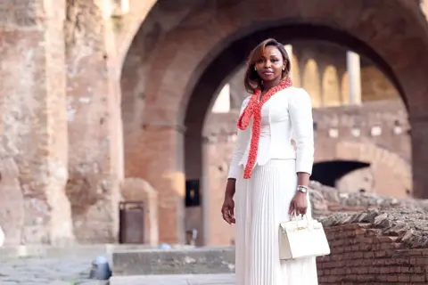 FRANCO ORIGLIA / GETTY IMAGES A woman dressed in white with a coral-coloured necklace stands inside a cloistered building in Rome.