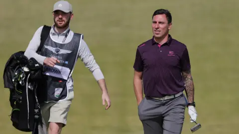 Getty Images Scott Brown, who is wearing a maroon polo shirt and dark grey shorts, walks along the fairway carrying his putter. A caddy, wearing a long sleeved grey top is walking to his right carrying a black golf bag. He is also wearing a silver cap. 