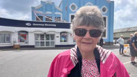 A woman with short grey hair, wearing sunglasses and a pink patterned T-shirt and pink coat stands in front of a blue and white Guernsey Electricity building.