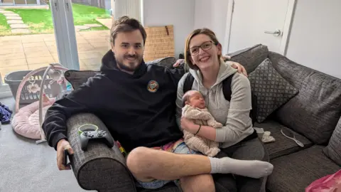 Sam, Emma and Sophie King are pictured together at home. They are sat together on a sofa. Emma is holding baby Sophie, who appears to be sleeping. The couple are smiling at the camera.
