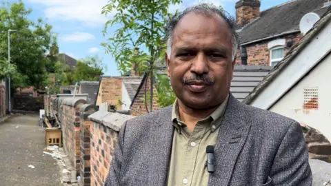 LDRS A man with grey hair at the sides of his head and a grey moustache stands in an alleyway behind a row of houses. He is wearing a grey suit jacket and a shirt.