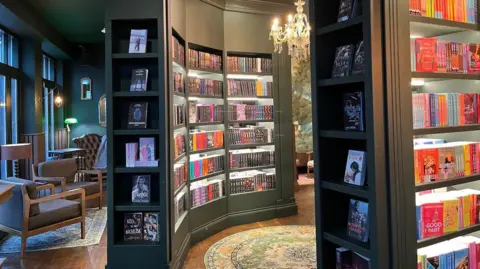 Alice Cullinane/BBC Two dark-green bookshelves are stocked with books and a green circular rug is placed on wooden floor. There are chairs placed by a window to the left. 
