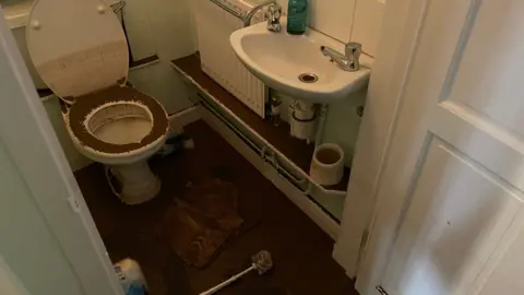A flood-damaged toilet. Brown mud is on the floor and is on the toilet seat itself. A toilet brush is non the floor. A sink and radiator are visible in the room.