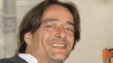Wayne Solomon has long brown hair and is smiling into the camera. He is wearing a shirt with a white collar and a grey jacket.