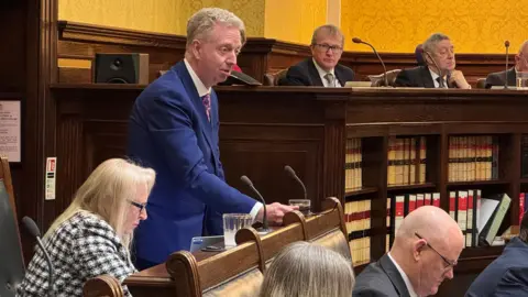 Alex Allinson standing in Tynwald while delivering his budget speech. He is wearing a blue suit, white shirt and red tie.